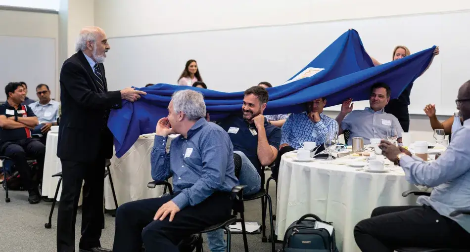 Principle-8_Example-1.jpg A photo of a role-playing event to educate the public on climate change issues. A man with white hair and a white beard wears a black suit and faces a group of people seated at tables. He holds one side of a large blue cloth that stretches over the heads of the people seated. A person stands behind the people at the tables and holds the other side of the cloth above their heads.
