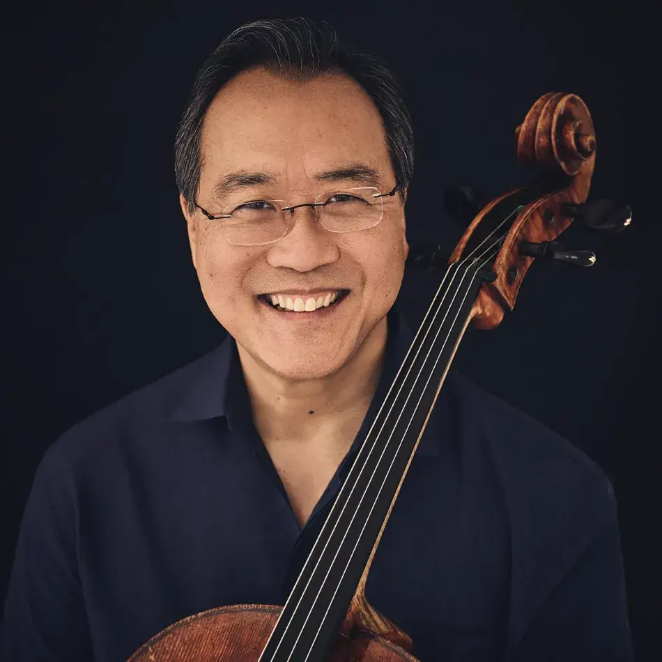 Photo by Jason Bell. A headshot of Yo-Yo Ma. He has brown skin and short gray hair, and wears a dark blue collared shirt. He holds a cello in his lap. Photo by Jason Bell.